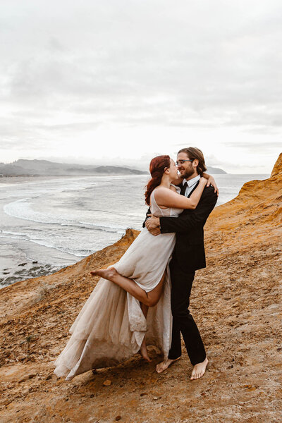 During their Oregon coast elopement, a couple dancces on top of a sand stone cliff  overlooking the occean. They are in their wedding attire and shoeless.