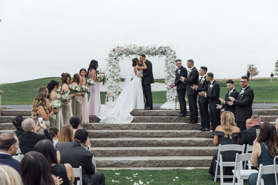 Trump National Golf Club | Wedding couple kissing under white floral arch on staircase with golf course backdrop | Bedminster, New Jersey