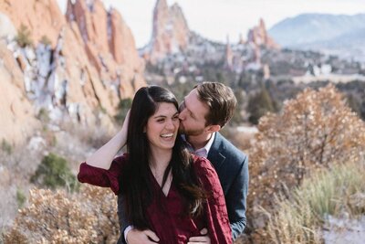 A man wraps his arms around his fiance and gives her a kiss on the cheek in front of red rock formations.