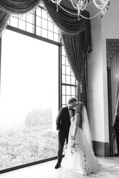 black and white image of a bride and groom standing in front of a floor to cieling picture window captured at the montecito club by santa barbara wedding photographer magnolia west photography