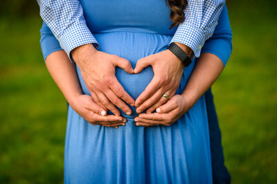 Close-up maternity photo of an expectant mother in a blue dress, with her partner's arms wrapped around her from behind, both creating a heart shape with their hands over her pregnant belly