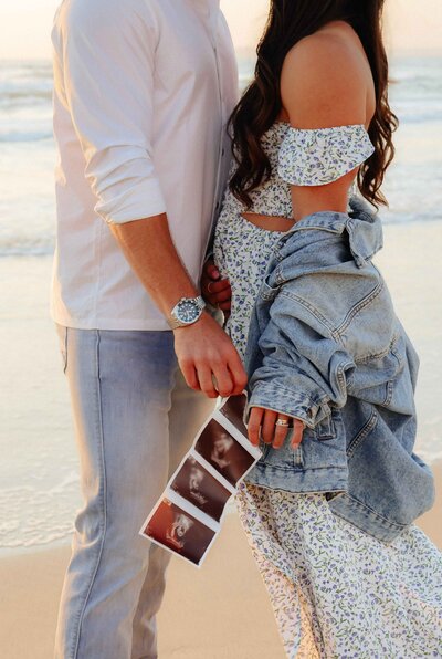 A happy pregnant couple stands on the beach, capturing a beautiful moment for their baby announcement in a lifestyle shoot.