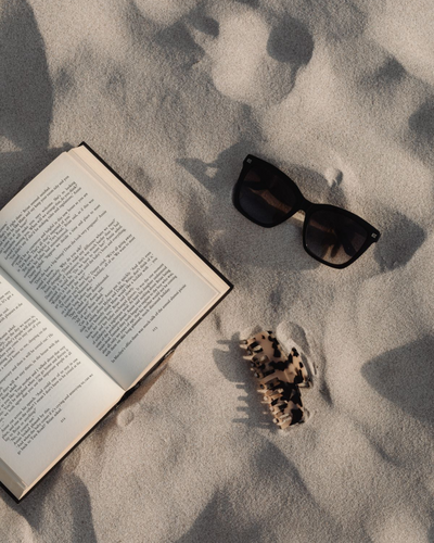 A book, black sunglasses, and a shell bracelet laid out on smooth sand with soft shadows