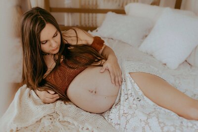 A pregnant woman with long hair lies on a bed, resting her hand on her bare belly and looking down at it, embodying the tender style of a Macon studio maternity photographer.