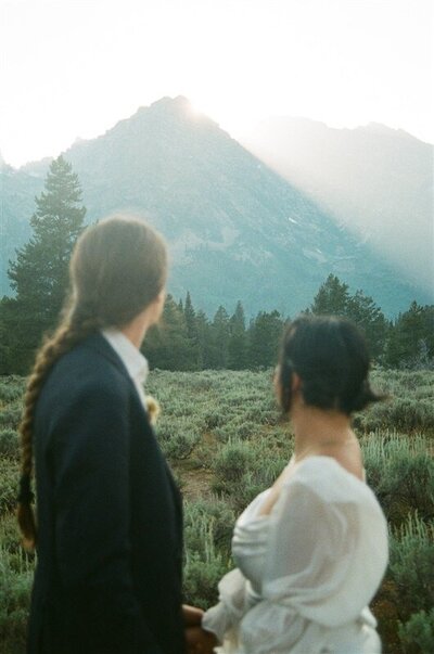 A couple stands in a field at sunset during their elopement in Jackson, Wyoming. They hold hands and look towards the mountains as the sun peeks over the peaks, casting a warm glow on the landscape.