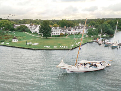 A sailboat glides across a calm river, with a picturesque shoreline in the background. White, colonial-style buildings are visible, surrounded by green trees and a manicured lawn dotted with white chairs. The sky is overcast.