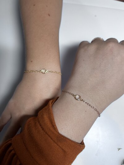 Two wrists with permanent jewelry there is a gold and silver chain and both have a charm attached.