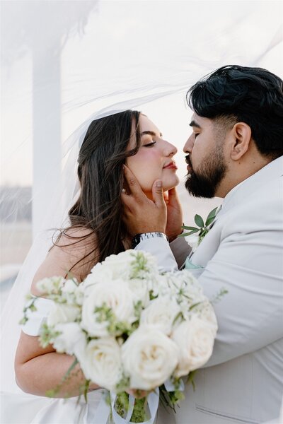 Bride and groom under a bridal veil kissing with a flower bouquet