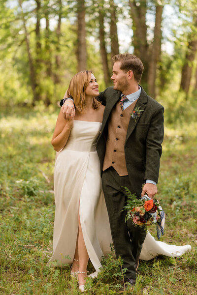 groom with his arm around his brides shoulder as he smiles at her and hold her bouquet