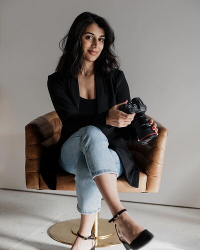 A person with long dark hair sits on a brown chair, holding a camera. They embody the New England lifestyle photographer aesthetic, wearing a black top, black sweater, and jeans, with legs crossed. The background is plain yet perfectly capturing the elegance of fine art weddings &amp; portraits in New England.