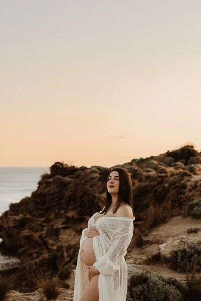 Pregnant mother with one arm leaning on her head and one hand holding her baby bump. Standing on a grass cliff with cliffs and ocean water behind her/ Hair flowing over her face as she closes her eyes and embraces her belly.