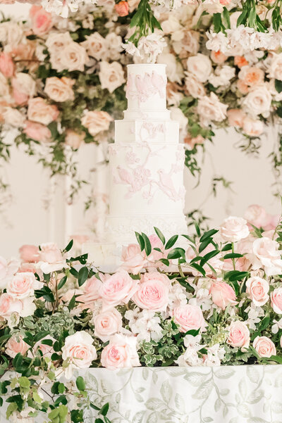 A gorgeous floral array of pink, white, and pastel florals on top of a warm green table cloth, fine china, and warm lighting surrounding a white wedding cake at Donovan Manor by North Carolina wedding photographer, JoLynn Photography