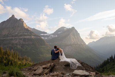 Bride and groom sit together on a rocky mountain overlook in Glacier National Park, Montana, embracing as the sun sets behind towering peaks. The bride’s veil flows in the wind, creating a breathtaking and intimate adventure elopement moment.