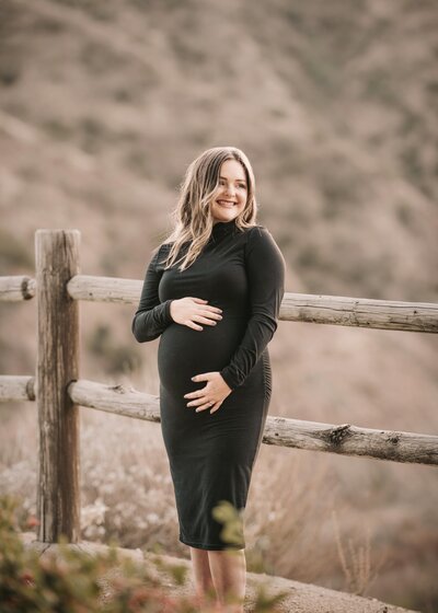 Expectant mom cuddling baby bump smiling in the distance. Elsinore mountains in the distance.