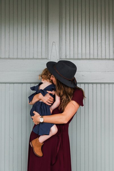 A woman in a burgundy dress and grey hat embraces a small child against a grey wooden background, perfecting her skills in family photography.
