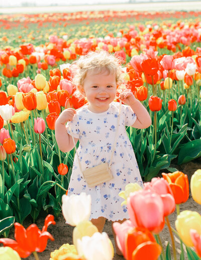 A little Dutch girl in a white dress with blonde curly hair smiles holding a little wooden camera.