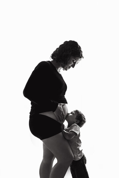 black and white photo of a maternity shoot. Mother wearing black long sleeve shirt and black shorts with toddler boy facing her and kissing her bump. Mother and son looking to each other while putting their hands on mother's bump.