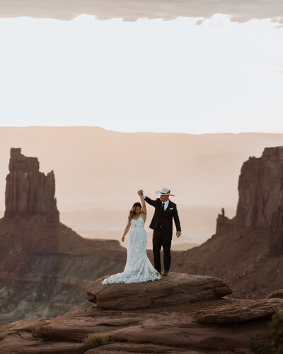 bride and groom holding hands in the air while standing on a rock formation in the desert