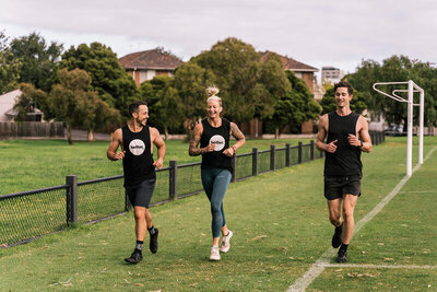 Three individuals running outdoors in a Melbourne park, wearing 'Project Better' branded tank tops, highlighting the gym's run club and sense of community."