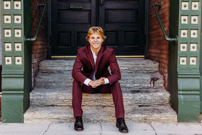 handsome young man sitting on steps in front of a black door while rubbing his hands together.