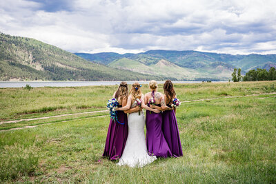 Bride and bridesmaids in purple dresses posing beside a peaceful mountain lake during a Colorado wedding.