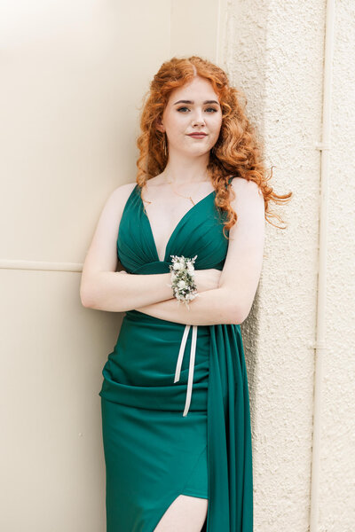 Red headed girl looking sassy in a stunning green dress, ready for her formal evening, standing against a cream coloured wall with her arms folded.