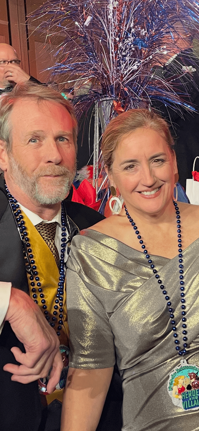 A man in a suit and a woman in a silver dress, The Nortons, both wearing blue bead necklaces, smile at an event with festive decorations.