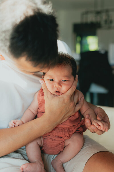 Photo of a dad holding his newborn baby so she is sitting up and he is cradling her head in his hands and her cheeks are all squishy taken by a langley photographer