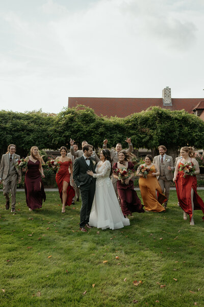 Bride and Groom are standing embracing each other as wedding party dressed in autumn color runs towards them in excitement