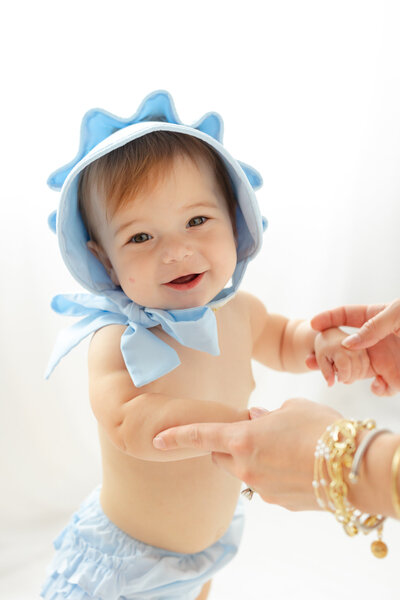 9 month old girl wearing a blue bonnet from TBBC Smiling at the camera holding her moms hand at a studio in Savannah