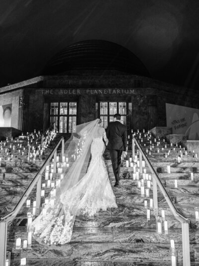 A bride in a flowing white gown and a groom in a classic black suit share a joyous moment together, posing in a lush garden setting captured in a timeless black and white photo.