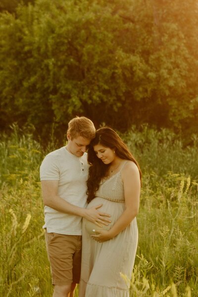 Husband and wife look down at the wife's expecting tummy in lush, open field.