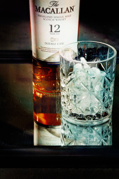 A refined image featuring a bottle of Macallan 12-Year-Old Scotch Whisky next to a crystal glass filled with ice. Captured by Mica McCook, this photograph emphasizes the elegance and sophistication of premium spirits.