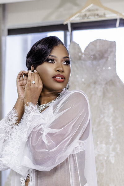 A glamorous bride in a luxury robe and red lipstick poses with her wedding dress in the background, radiating elegance and anticipation on her special day. A timeless bridal preparation moment.