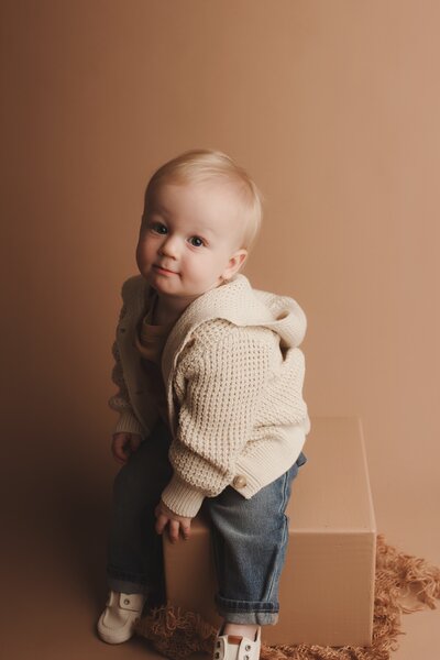 1 year old sitting on a tan box on a tan backdrop smiling
