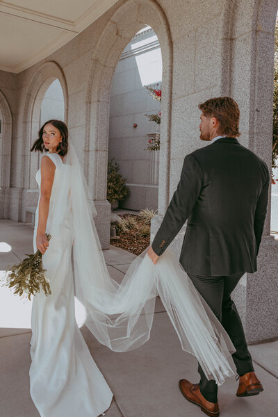 A groom walks behind his bride while holding her long flowy vail at [venue name] venue