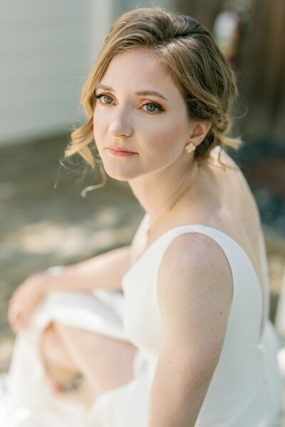 Kohnur Bride with soft glam makeup featuring warm peach eyeshadow, fluttery lashes, natural brows, and nude pink lips, styled in a romantic low bun wearing a minimalist white gown at an intimate garden wedding.