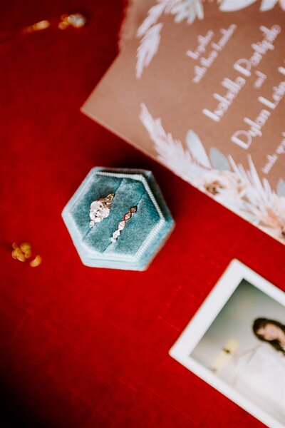 An elegant wedding detail shot on a red surface featuring a teal velvet ring box open to reveal a delicate bridal hairpiece. The background includes a blurred wedding book with the names 'Isabella & Dylan' and a picture frame showing a soft-focused image of the bride.