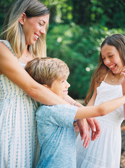 Mother and two tween children laugh joyously amidst green summer setting.
