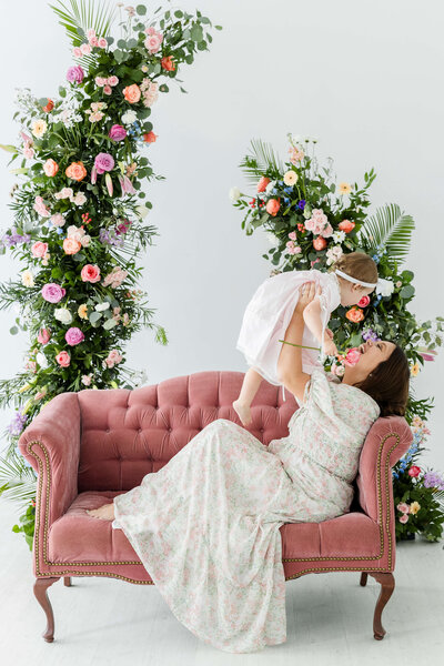 Mom holding baby up while sitting on couch surrounded by flowers