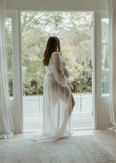 Pregnant mama cradling her baby in a see-through white robe in doorway to backyard