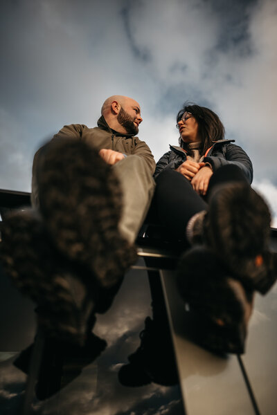 Low-angle photo of a wedding photographer and videographer duo sitting together on a ledge, looking at each other with the sky above. Based in the Lake District, UK, and capturing destination weddings worldwide