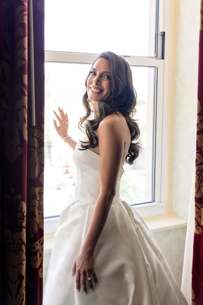 A woman in a white wedding dress stands by a window, smiling over her shoulder. She has long, wavy hair and is surrounded by floral-patterned curtains. Sunlight brightens the room, highlighting her joyful expression.