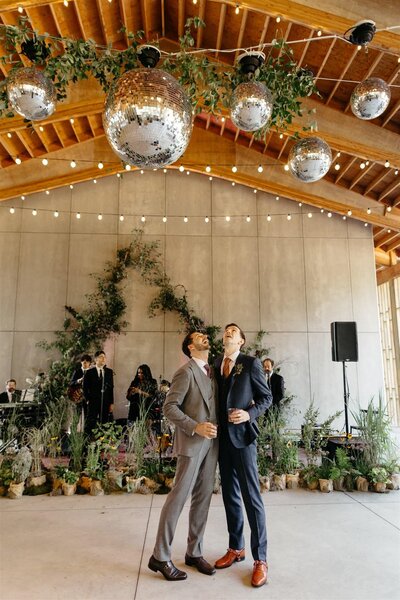 Two dapper grooms admire the disco balls and twinkle lights above. Botanical focal point behind them, with dozens of potted plants.