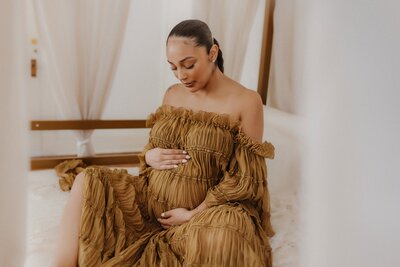 A woman in an off-shoulder, ruffled brown dress sits on a bed, gently cradling her pregnant belly with both hands—a serene moment captured by a Macon maternity photographer.