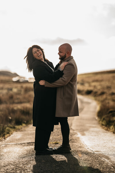 A joyful wedding photographer and videographer duo laughing and embracing on a countryside path in the Lake District, UK. Specialists in capturing weddings across the Lake District and destinations worldwide.