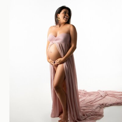 Pregnant first time mom to be Gabriella of columbian decent laughing and looking over her shoulder while holding her bare pregnant belly wearing a pink open front long flowy dress studio portrait created in Fort Mill SC