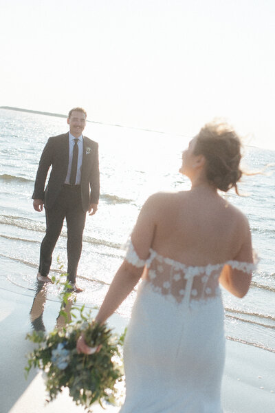 bride and groom walk toward each other on sandy beach with bright sun behind
