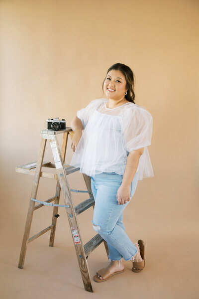 Monique Anne smiling next to a Pentax camera on a ladder in a studio