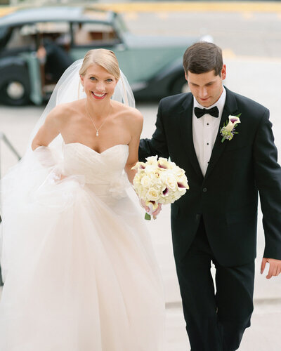 A couple posing for their wedding portrait in their wedding gown and tux walking up steps groom assisting her up the steps with antique car in the backgound.